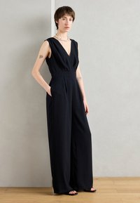 Svart ermeløs jumpsuit med dyp V-hals, brede ben og sidelommer. Stoffet ser glatt og lett ut.