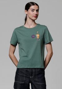 Kurzes grünes Baumwoll-T-Shirt mit Rundhalsausschnitt, das ein Cartoon-Katzendesign und Text zeigt, kombiniert mit dunkelblauen Jeans.