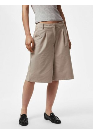 Person iført beige knælange vidde shorts, grå ærmeløs top og sorte loafers, stående på en ensfarvet hvid baggrund.
