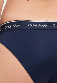 Námornícky modré bikiny s hladkou textúrou, s širokým elastickým pásom označeným "Calvin Klein" v bielej farbe.