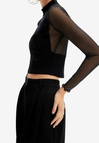 Musta lyhyt toppi, jossa on mock neck, ribbetty rakenne ja läpikuultavat pitsihihat. Yhdistettynä korkeavyötäröisiin mustiin housuihin, joissa on erityinen vyötärönauha.
