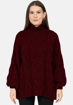 Woman wearing a dark red, cable-knit turtleneck sweater with balloon sleeves and black pants on a light background.