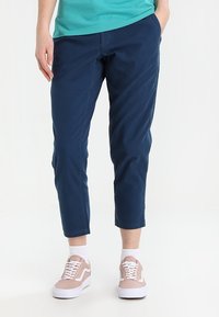 Pantaloni cropped blu navy in tessuto liscio con tasche laterali e dettagli di cucitura discreti, abbinati a sneakers rosa chiaro e calze bianche.