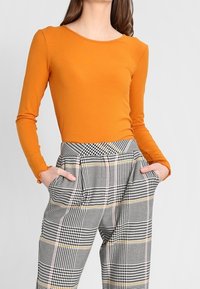 Frau trägt ein orangefarbenes langärmliges Rippoberteil und hochtaillierte schwarz-weiß-gelb karierte Hosen mit den Händen in den Taschen.