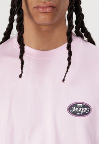 Camiseta de algodón rosa claro con un parche de logo redondo que incluye texto e ilustración, con cuello redondo y mangas cortas.