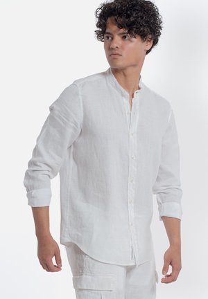 Giovane uomo con capelli ricci che indossa una camicia di lino bianca a maniche lunghe con maniche arrotolate e pantaloni cargo bianchi, guarda di lato.