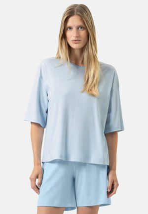 Blonde vrouw die een loszittende lichtblauwe top met korte mouwen en subtiele strepen draagt, gecombineerd met effen lichtblauwe shorts, staande tegen een witte achtergrond.