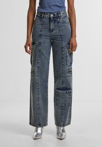 Cargo jeans in faded blue denim, wide-leg fit with multiple pockets, and silver hardware accents. Paired with metallic silver shoes.