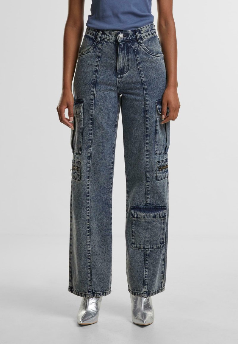 Cargo jeans in faded blue denim, wide-leg fit with multiple pockets, and silver hardware accents. Paired with metallic silver shoes.