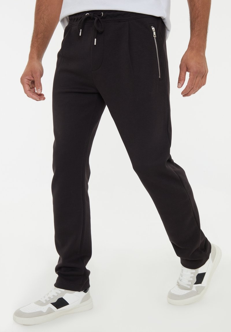 Homme portant un jogger noir avec des poches zippées et des baskets blanches, debout jambes écartées sur un fond uni.