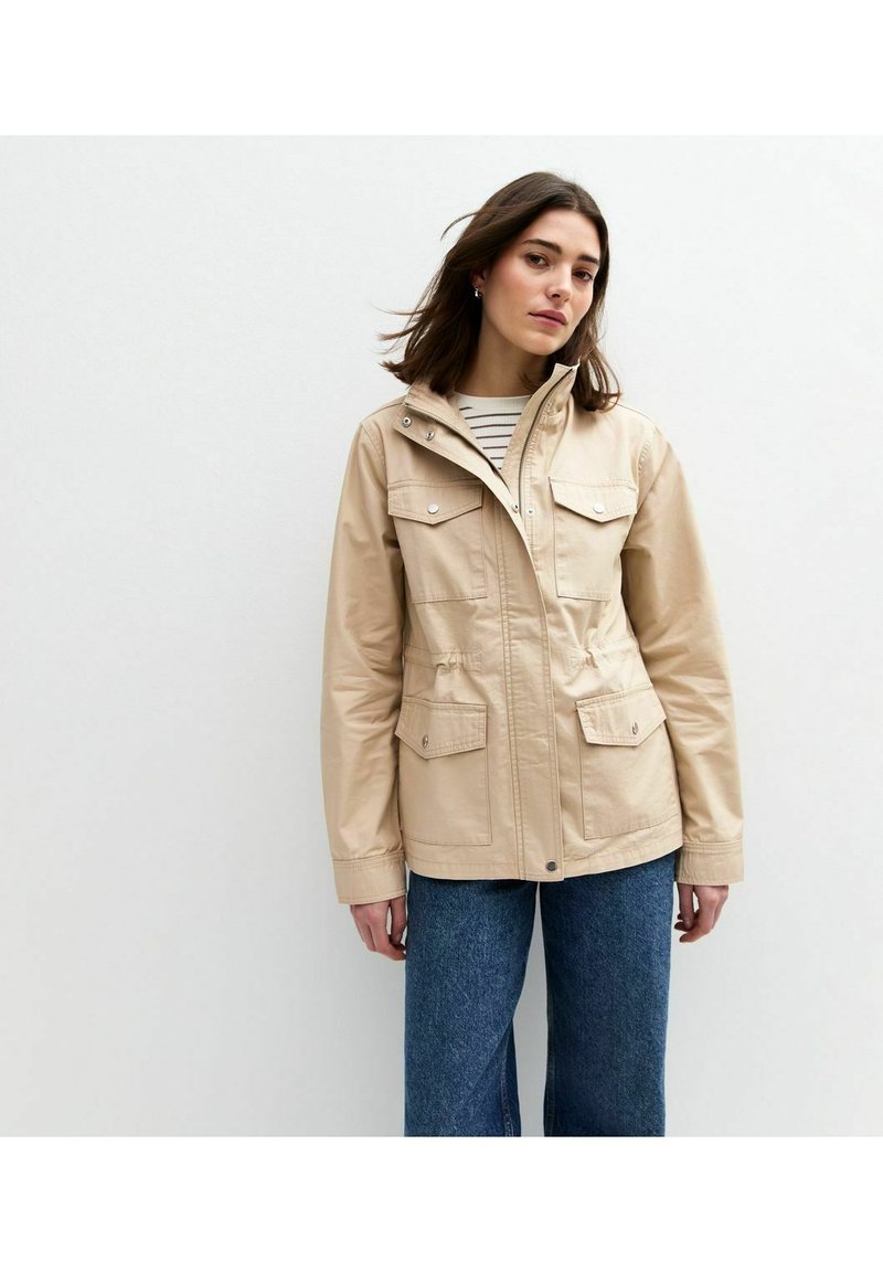 Beige jas met een ritssluiting en drukknoopsluiting, twee voorzakken en een afneembare capuchon, gedragen over een gestreept shirt en blauwe jeans.
