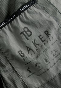 Šedá tkanina s mriežkovaným vzorom, so štítkom "BAKER by TED BAKER." Ikony naznačujú odolnosť voči vode, trvanlivosť, teplo a recyklovanú výplň.