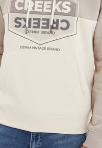 Sudadera de color beige claro con un bolsillo frontal, cuello con cordón y texto gráfico en gris oscuro. Tejido suave con puños y dobladillo acanalados.
