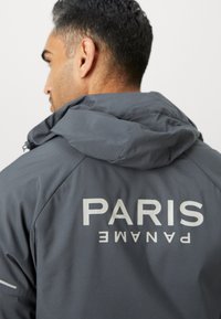 Chaqueta con capucha gris que presenta el texto reflectante "PARIS PANAME" en la parte posterior, hecha de tela suave con un acabado ligero y texturizado.