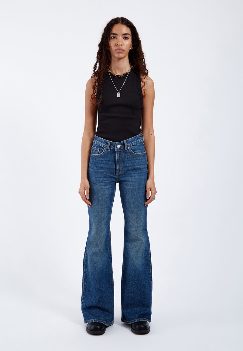 Woman with long curly hair wearing a sleeveless black top, high-waisted blue flared jeans, black shoes, and silver necklaces against a white background.