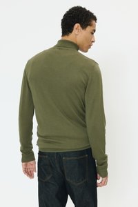 Oljčno zelena pulover z dolgimi rokavi in visokim ovratnikom, narejena iz mehke tkanine. Prilegajoč kroj, nošena s temno modrimi jeans hlačami.