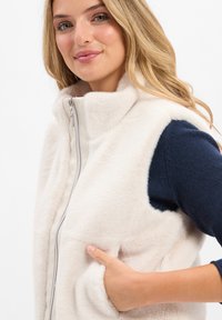 Fleece vest in cream with a high collar, front zipper, and side pockets; soft texture and sleeveless design complemented by navy shirt.