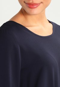 Blusa blu navy con scollatura rotonda e texture liscia, con dettagli di cucitura discreti lungo il colletto e le spalle.
