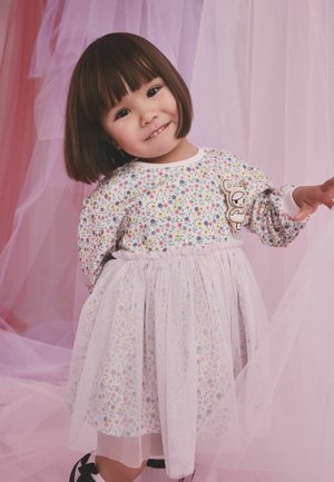 Mladi djevojčica s kratkom tamnom kosom u cvjetnoj haljini s til suknjom, smiješi se i pruža ruku nasuprot meke ružičaste tkanine.