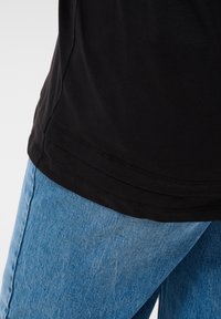 Gros plan sur la partie inférieure d'une chemise noire portée sur un jean en denim bleu clair, sur un fond gris clair.