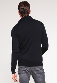 Svart stickad tröja med en åtsittande design och en krage. Har långa ärmar och ribbade muddarna, kombinerat med ljusgrå jeans.