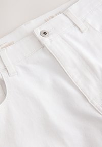Jean slim fit blanc en denim en gros plan montrant le bouton, les passants de ceinture et les détails de la poche avant.