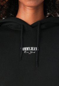 Sweatshirt preto com cordão, apresentando um logotipo branco "TOMMY JEANS New York" no peito. Textura suave com um ajuste justo.