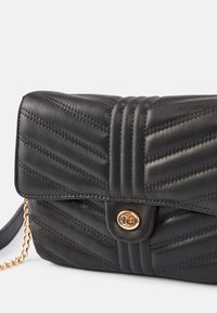 Zwarte doorgestikte leren crossbodytas met gouden draaivergrendeling en gouden kettingriem, getoond tegen een witte achtergrond.