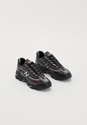 Paire de chaussures de sport New Balance noires et grises avec des détails rouges et des semelles épaisses sur fond blanc.