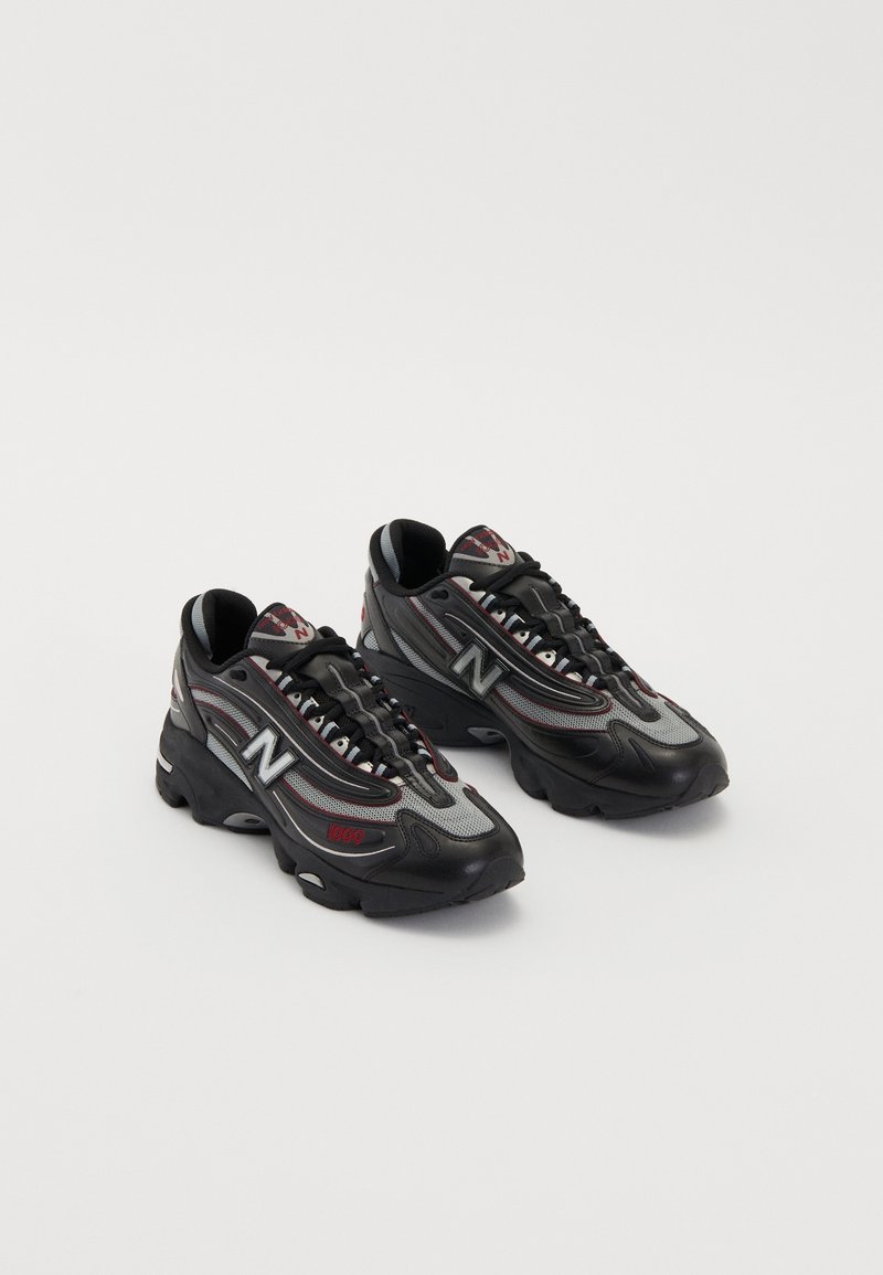 Paire de chaussures de sport New Balance noires et grises avec des détails rouges et des semelles épaisses sur fond blanc.