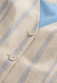Ľanová košeľa s krémovým základom a svetlomodrými pruhmi, vybavená kruhovými béžovými gombíkmi a hladkou, kontrastnou modrou látkou vo vnútri.