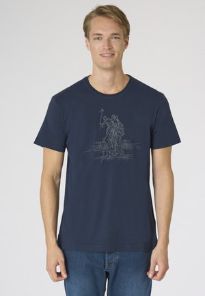 Giovane uomo con una T-shirt blu navy con un giocatore di polo a cavallo disegnato e il logo USPA, in piedi davanti a uno sfondo chiaro e uniforme.