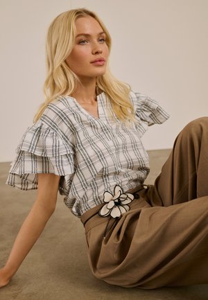 Lyshåret kvinde sidder på gulvet iført en flæset ternet bluse og brune bukser med et blomstret bælteaccessory.