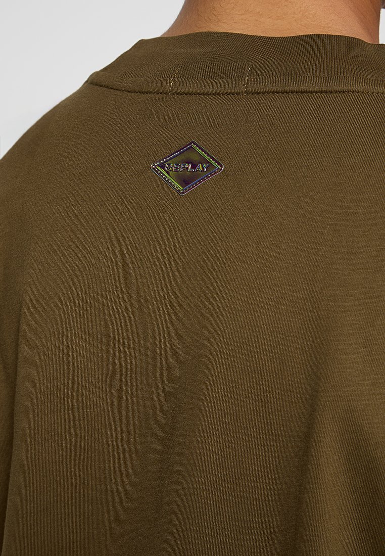 Olivengrünes Baumwoll-T-Shirt mit runder Ausschnitt und einem strukturierten, diamantförmigen Logo-Patch auf der Rückseite, das mehrfarbige Akzente aufweist.