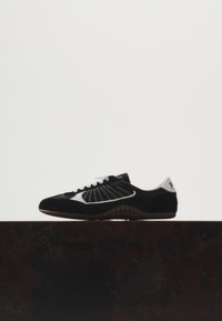 Baskets noires et blanches avec des lacets blancs sur une surface sombre, montrant le profil latéral contre un fond clair uni.