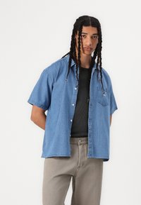 Short-sleeve denim shirt in light blue. Features a left chest pocket and button-up design. Worn over a black ribbed tank top and light gray pants.