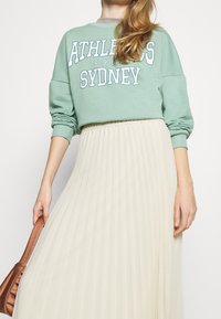 Mintgrön sweatshirt med vit text, tillsammans med en plisserad krämfärgad midi-kjol. Modellen håller en brun väska.