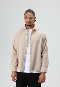 Lichtbeige overhemd met knopen, gemaakt van zachte stof, met een brede kraag en lange mouwen. Gedragen over een wit T-shirt. Zwarte broek zichtbaar.