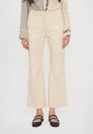 Femme portant un pantalon évasé couleur crème, des chaussures plates noires avec des boucles dorées et un haut à manches longues avec des motifs floraux et géométriques.
