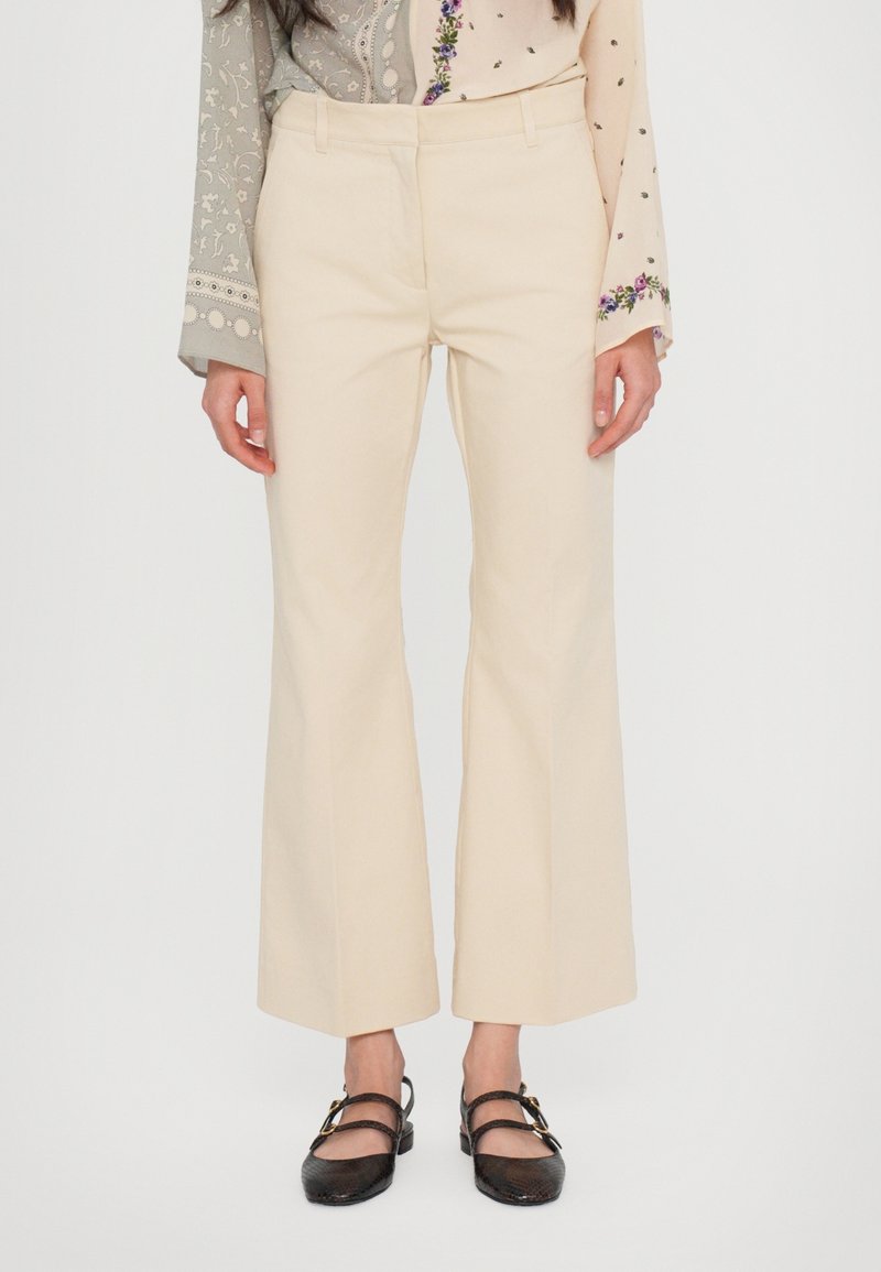Femme portant un pantalon évasé couleur crème, des chaussures plates noires avec des boucles dorées et un haut à manches longues avec des motifs floraux et géométriques.