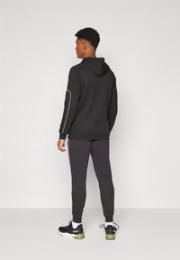 Czarna bluza z kapturem z regulowanym kapturem, z białymi akcentami po bokach, w zestawie z ciemnoszarymi dresami. Model ma na sobie czarne sneakersy z zielonymi akcentami.
