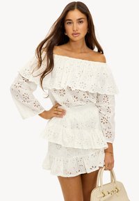 Jeune femme aux longs cheveux portant une robe blanche à fleurs avec épaules dénudées en tissu ajouré, tenant un sac à main beige, debout avec une main sur la hanche.