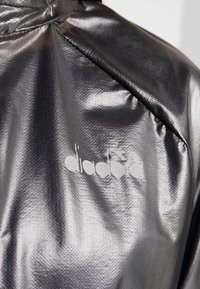 Tissu gris métallisé brillant avec une couture diagonale et un logo Diadora blanc délavé sur un vêtement.