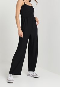 Žena v černém bezramínkovém overal s širokými nohavicemi a bílými vysokými teniskami, stojící na pozadí v jemně světle barevném pozadí.