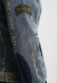 Rękaw dżinsowej kurtki z naszywką "Ranger Challenge", widoczne zużycie i ciemnoniebieska łatka na łokciu.