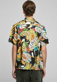 Chemise à manches courtes arborant un motif tropical vif de bananes et de feuilles sur un fond noir. Le matériau semble léger et lisse.