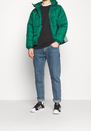 Doudoune verte à capuche, fermeture éclair et poignets élastiques, associée à un t-shirt noir et un jean bleu, complétée par des baskets noires et blanches.