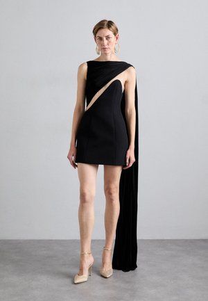 Black asymmetrical dress with draped fabric, showcasing a side cutout. Features a fitted bodice and floor-length cape. Gold embellished heels.