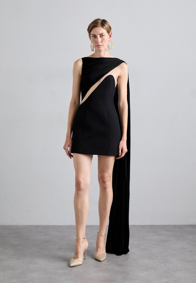 Juoda asimetrinė suknelė su drapiruota medžiaga, demonstruojanti šoninį išpjovą. Suknelė turi prisiūtą liemenį ir grindis siekiančią kampinę. Auksu dekoruoti aukštakulniai.