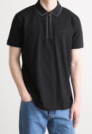 Jeune personne portant un polo noir manches courtes avec une fermeture éclair à l'avant et un jean bleu, main dans la poche, debout devant un fond uni.